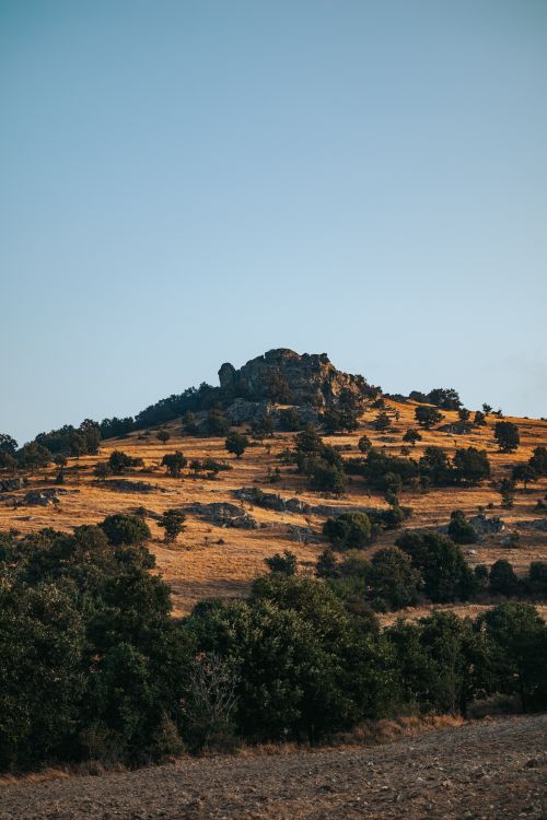 Free Beautiful countryside landscape with a rocky hill and scattered trees under a clear blue sky. Stock Photo