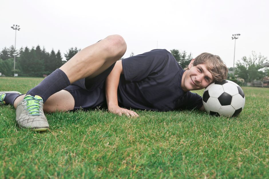 Teen soccer player smiling on grass field resting with a soccer ball.