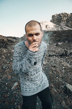 A stylish man with a bald head smokes a cigarette in a rocky mountain landscape.
