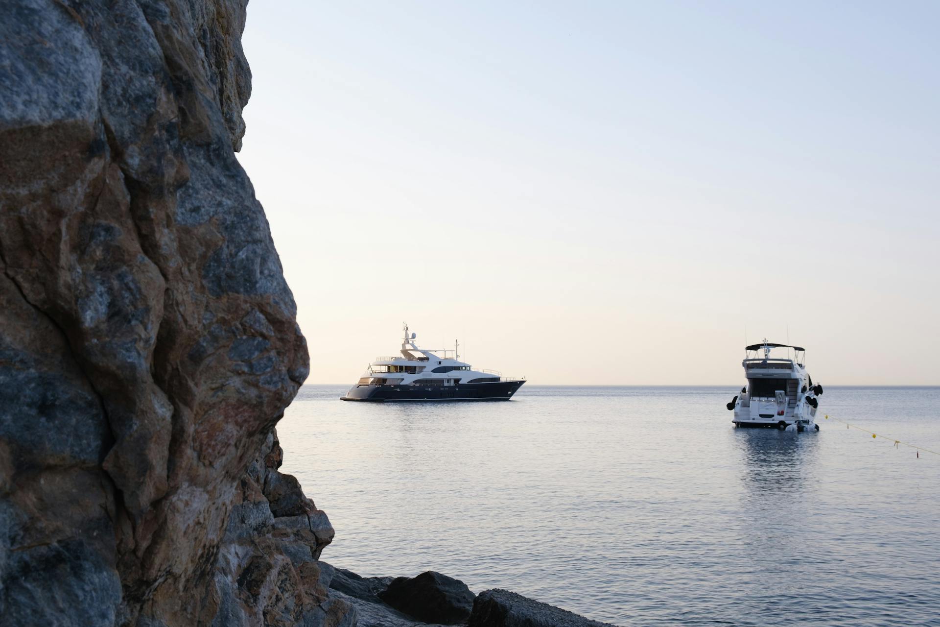 Two luxury yachts anchored near a rocky shoreline during a serene sunset.