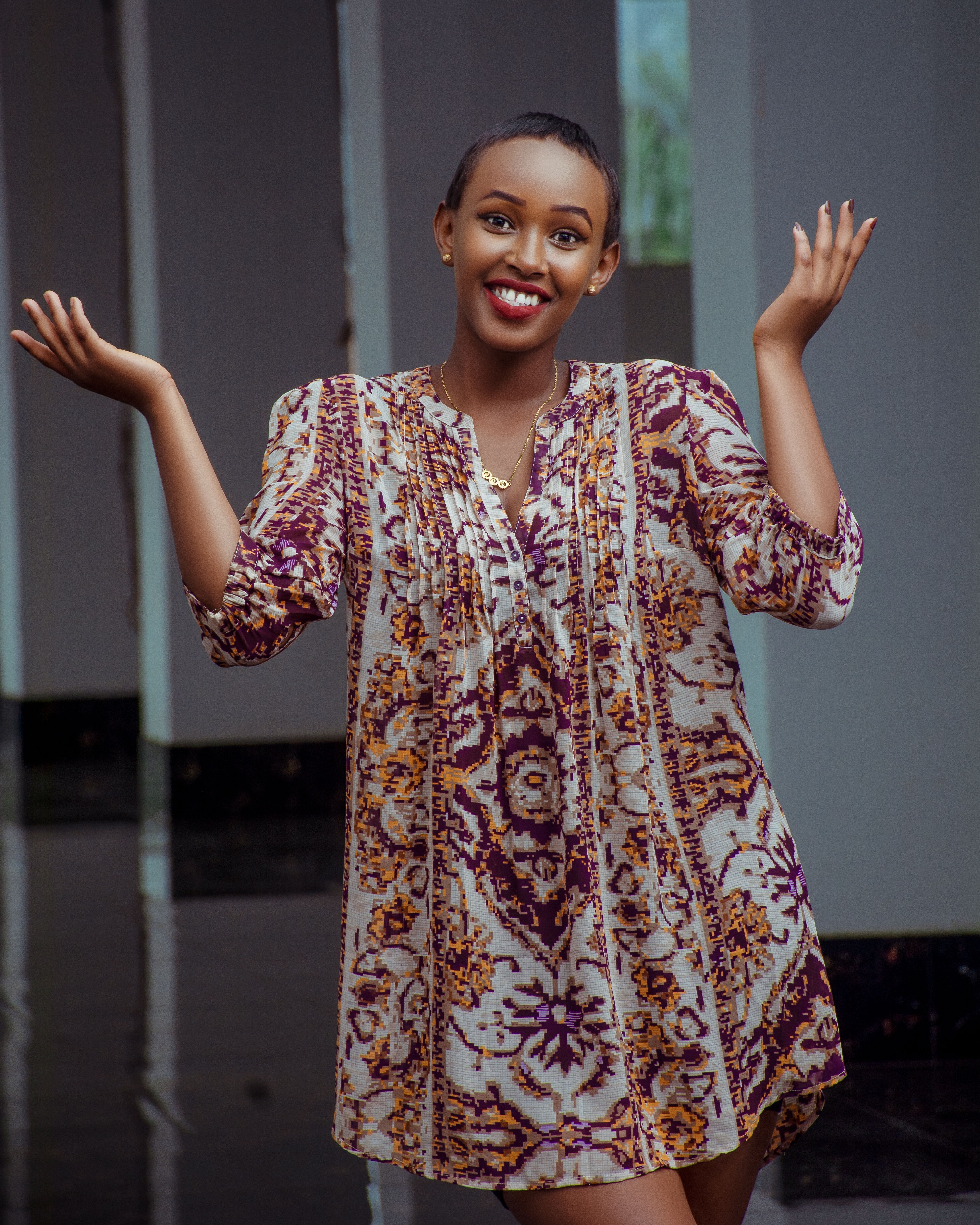A cheerful African woman in a floral dress stands indoors, radiating happiness with a big smile.