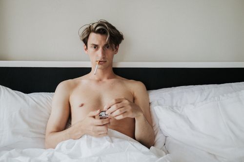 Free Shirtless man sitting on white bed holding a lighter with a cigarette. Stock Photo