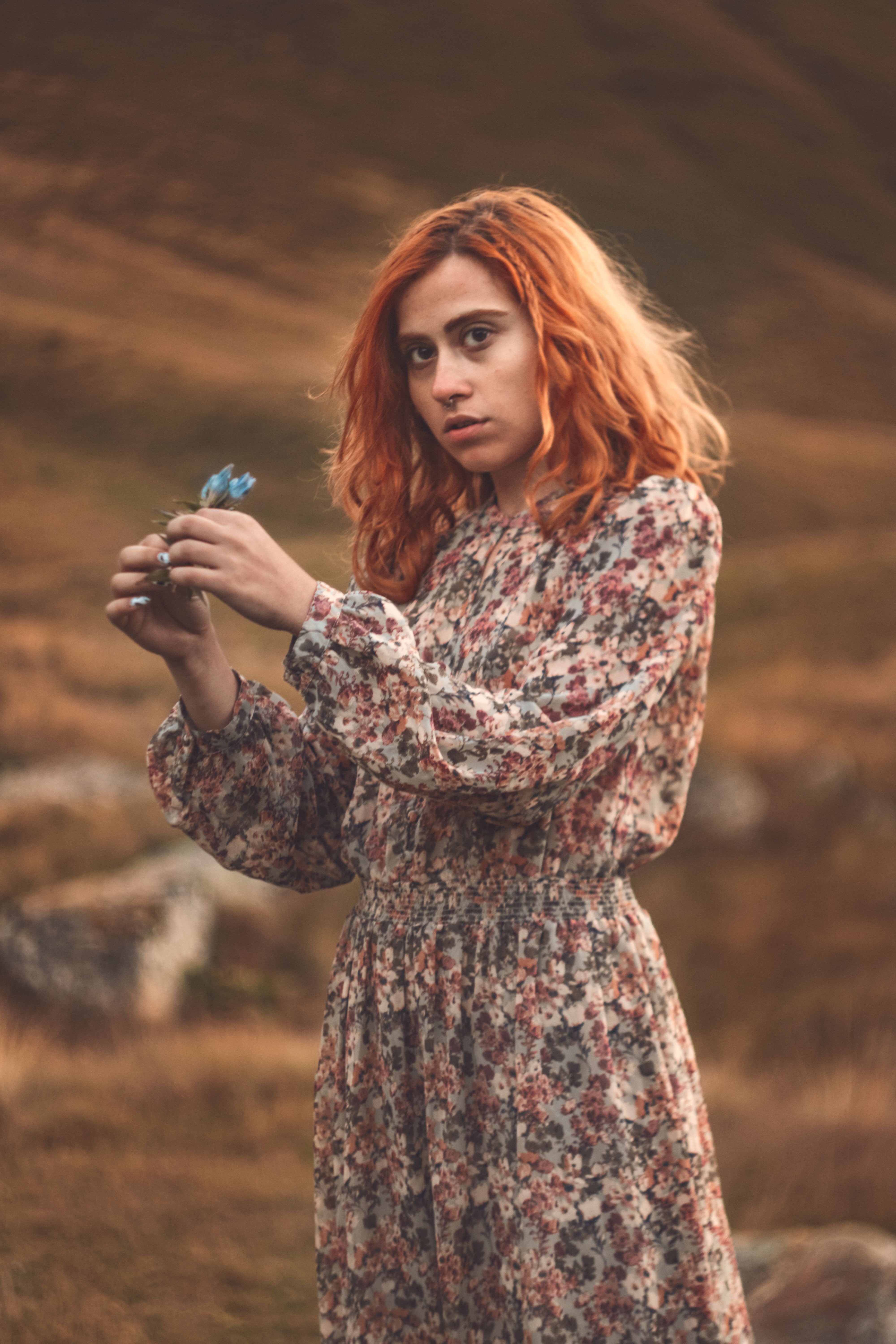 A young woman with red hair in a floral dress holds a blue flower in an outdoor setting, exuding a vintage vibe.