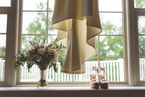 Flower Bouquet and Shoes on Heels Standing on Windowsill Beside Hanging Wedding Dress 