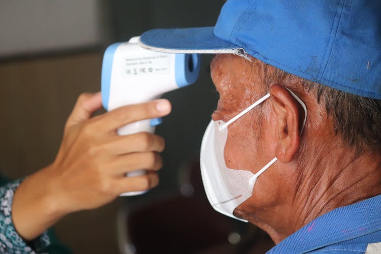 Person Measuring Temperature Of Old Man In Blue Cap By Thermometer Gun 