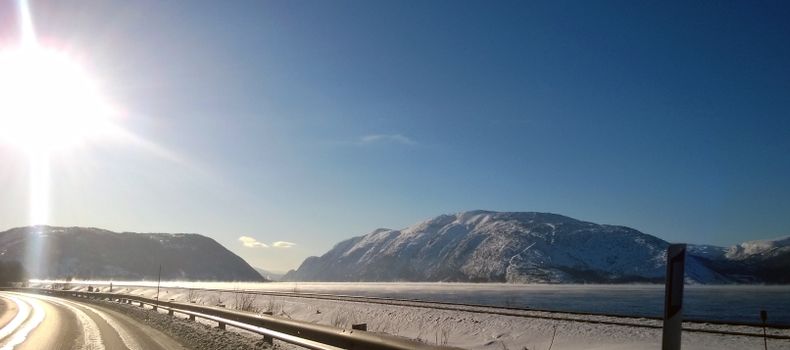 A sunlit winter road winding through snowy mountains in Hol, Norway, offering a scenic view.
