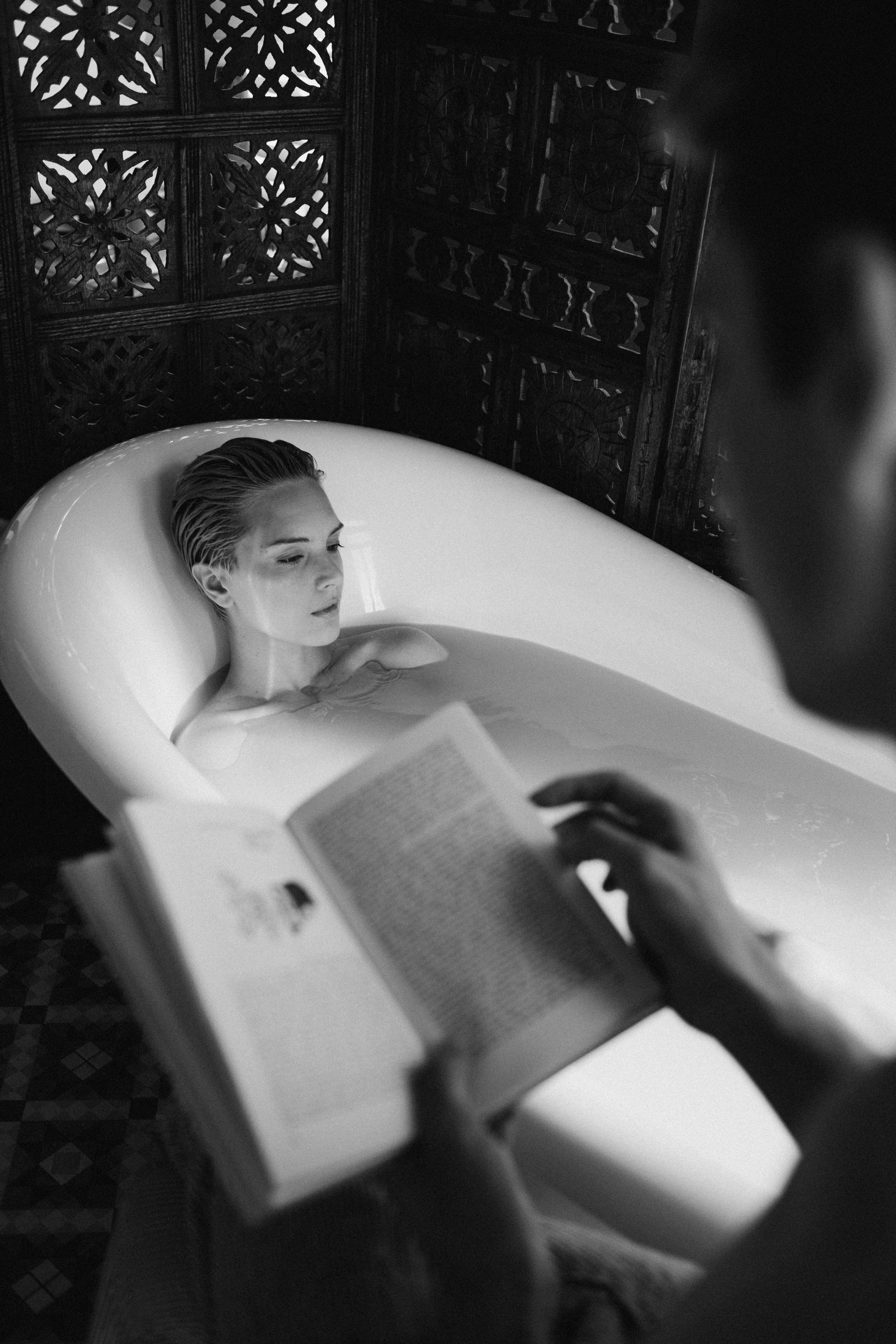 A young woman relaxes in a bathtub reading a book, captured in black and white photography.
