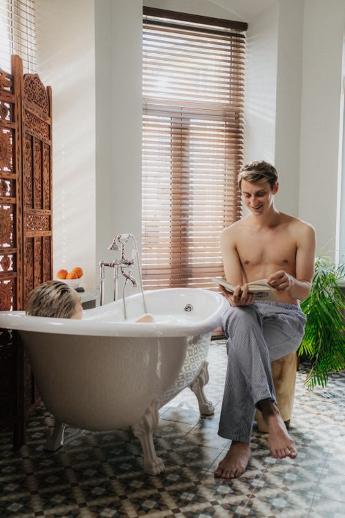 Free A man reading a book beside a bathtub in a sunlit room, creating a peaceful atmosphere. Stock Photo