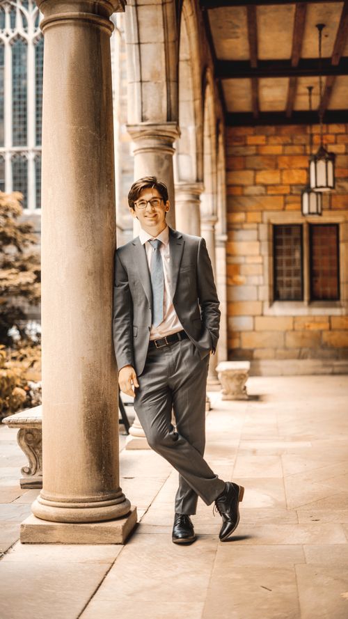 Free Smartly dressed man in a suit leaning against a pillar in an elegant arched corridor, smiling. Stock Photo