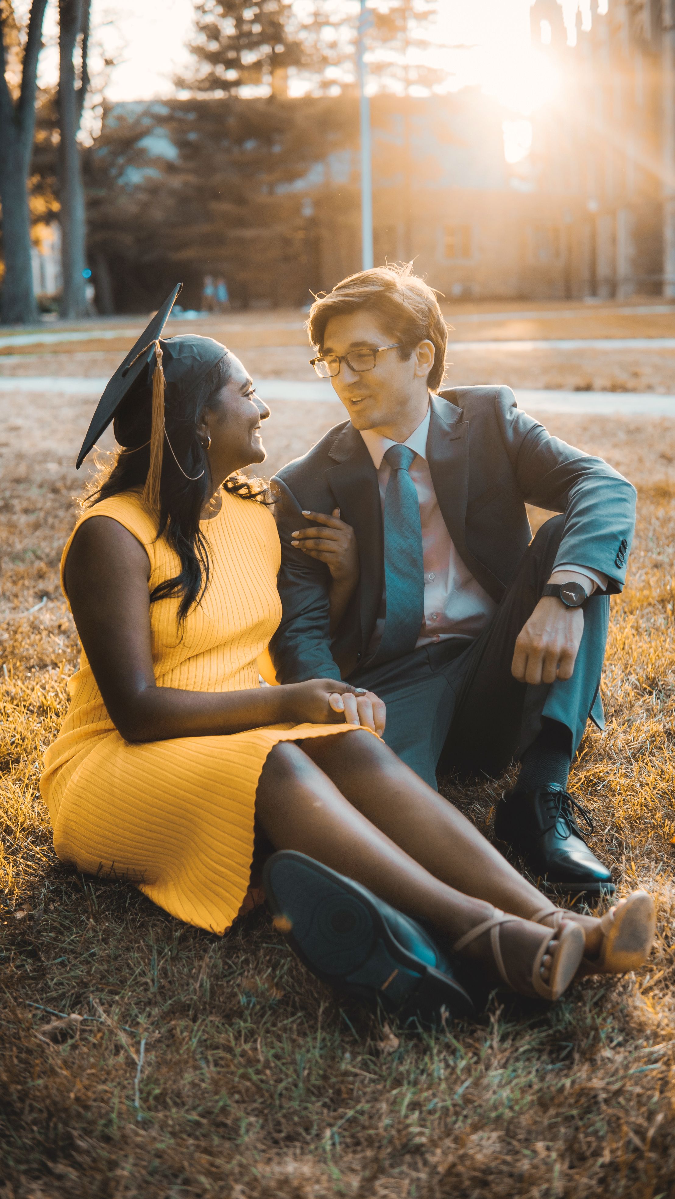 Interracial couple celebrating graduation in a sunny park, sitting on grass, sharing a joyful moment.