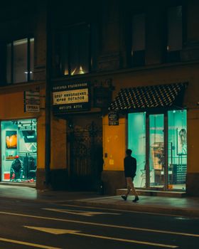 Man walking past illuminated shops on a city street at night.