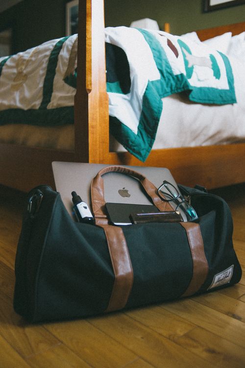 Free Cozy bedroom with a travel bag, laptop, and accessories on wooden floor, ideal for lifestyle imagery. Stock Photo