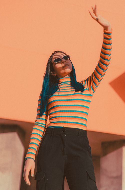 Free Trendy teenager with sunglasses poses confidently in a colorful striped outfit against an orange backdrop. Stock Photo