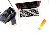 白色床上的时尚生活必需品包括笔记本电脑、手提包、手机和香水