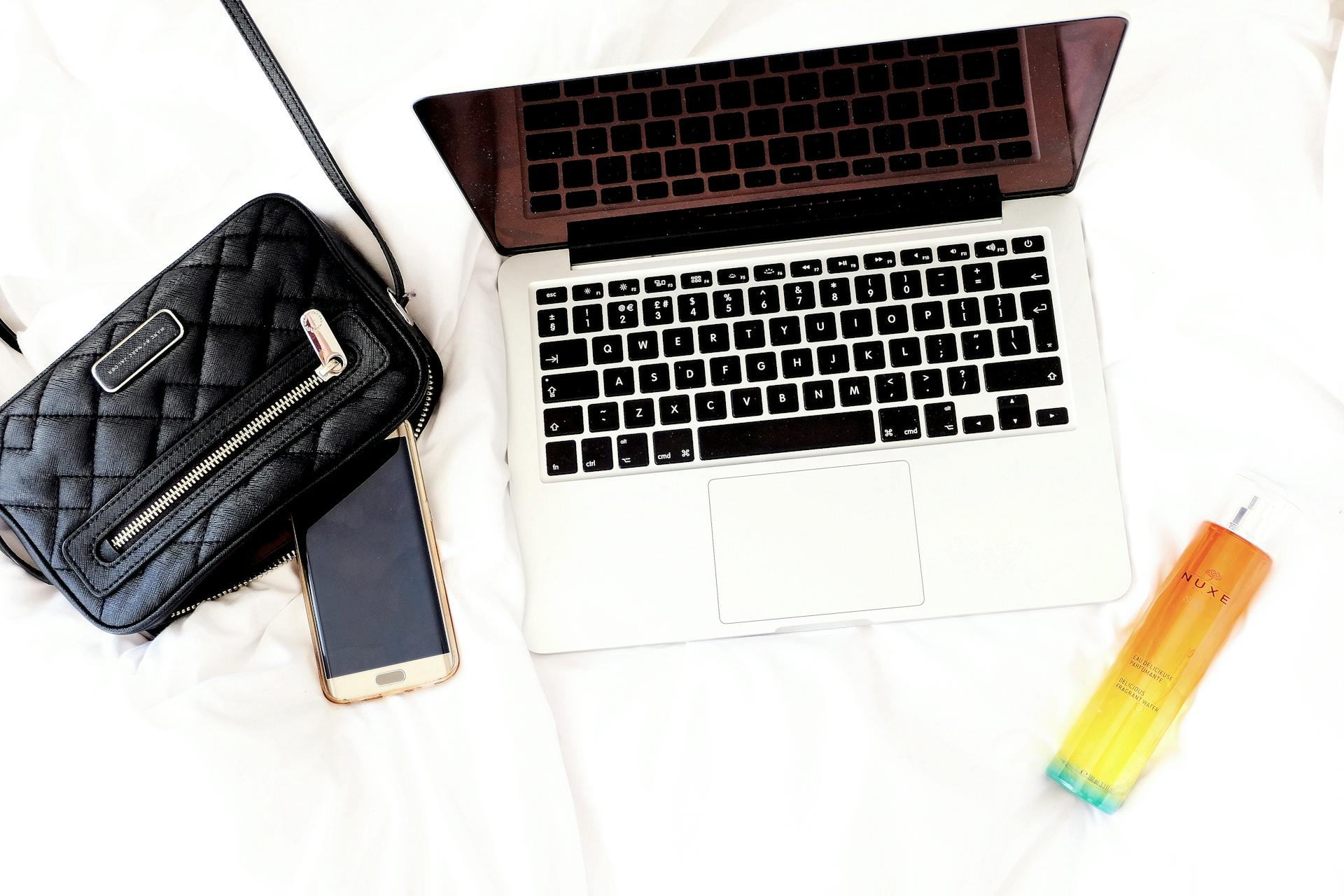 Stylish lifestyle essentials including a laptop, handbag, phone, and perfume placed on a white bed.