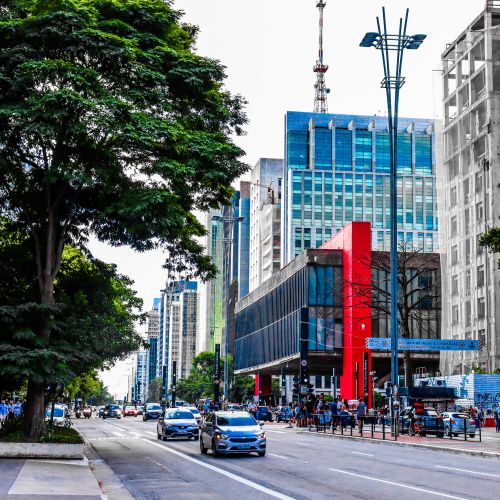 Free Lively street scene featuring modern high-rise buildings, cars, and lush trees in an urban setting. Stock Photo