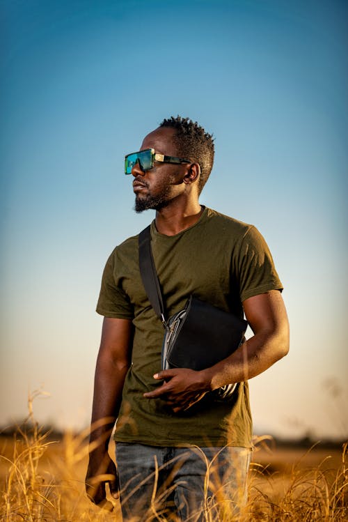 Free Man Standing on Grass Field while Wearing Sunglasses and Carrying His Sling Bag Stock Photo