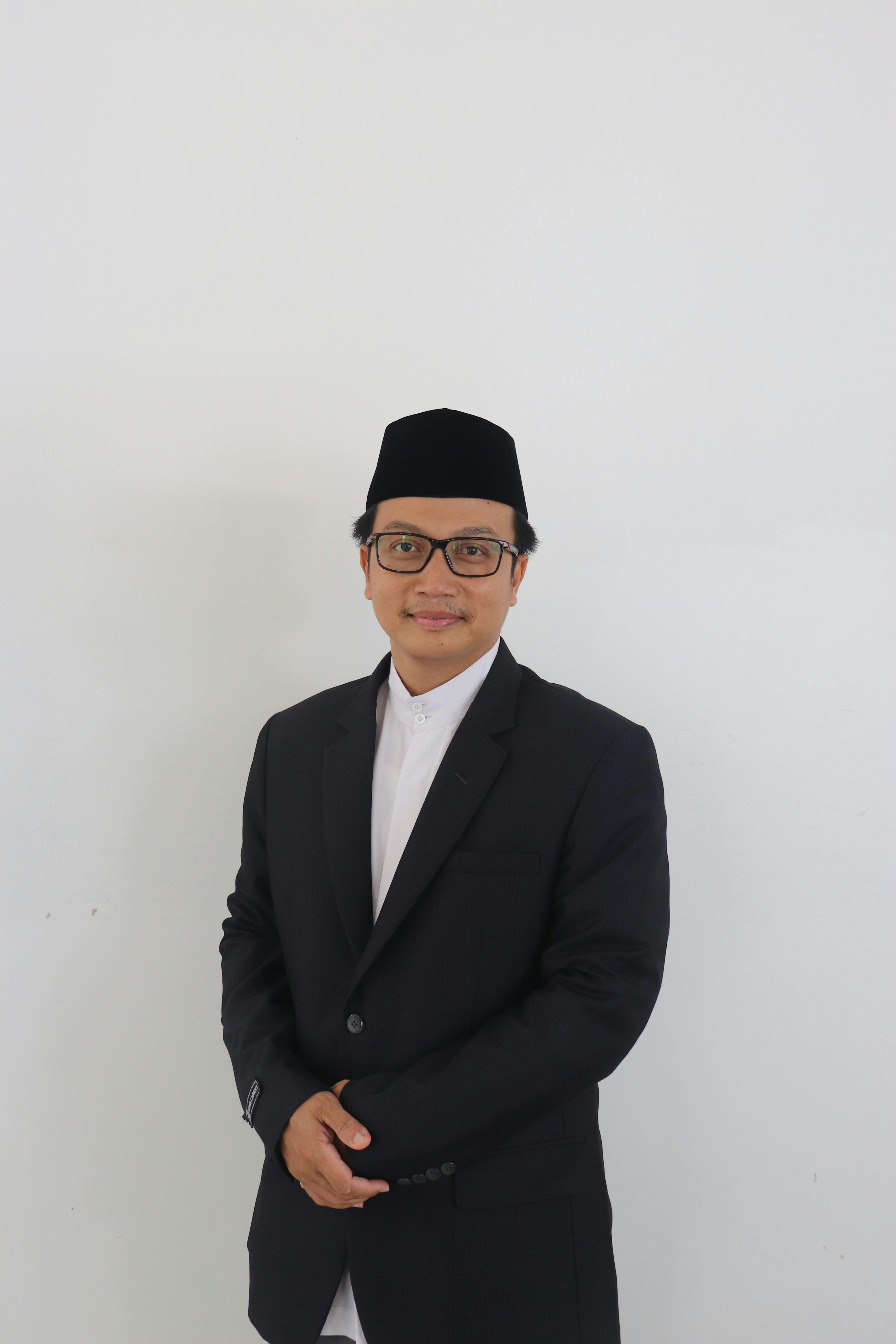 Portrait of a man wearing glasses, a black suit, and taqiyah, standing confidently against a white background.