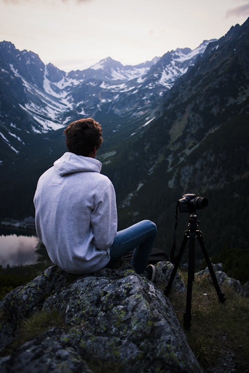 Uomo In Felpa Con Cappuccio Grigia Che Si Siede Sulla Pietra Grigia Con Treppiede Della Fotocamera Dslr