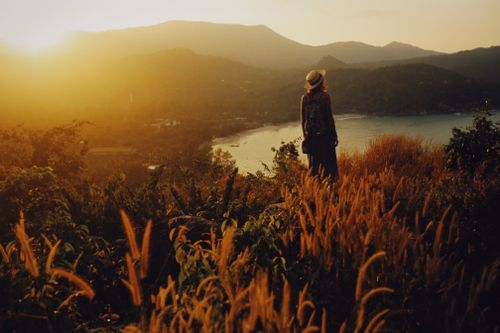 Free A woman in a hat stands on a hilltop at sunset, gazing over lush hills and a serene coastline. Stock Photo