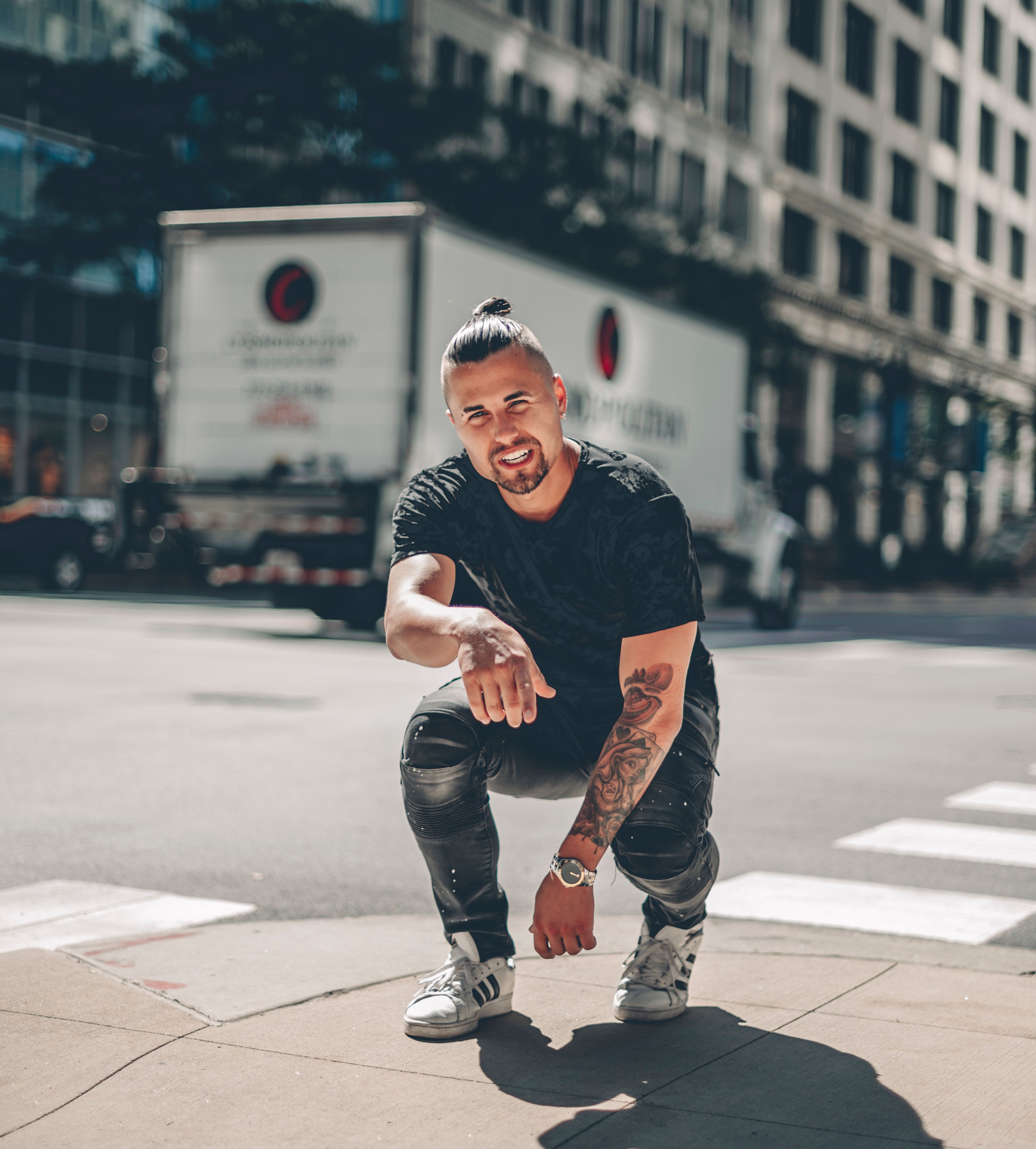A fashionable young man with tattoos poses confidently on a sunlit urban street corner.