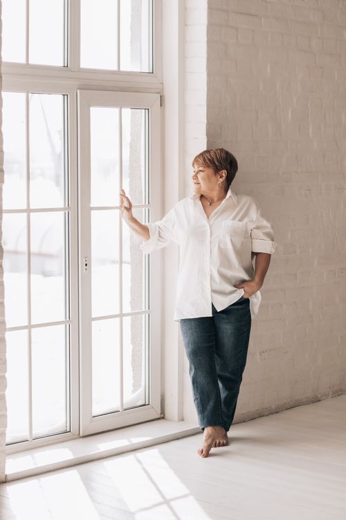 Free Elegant middle-aged woman in casual attire gazing through a sunlit window in a serene indoor setting. Stock Photo