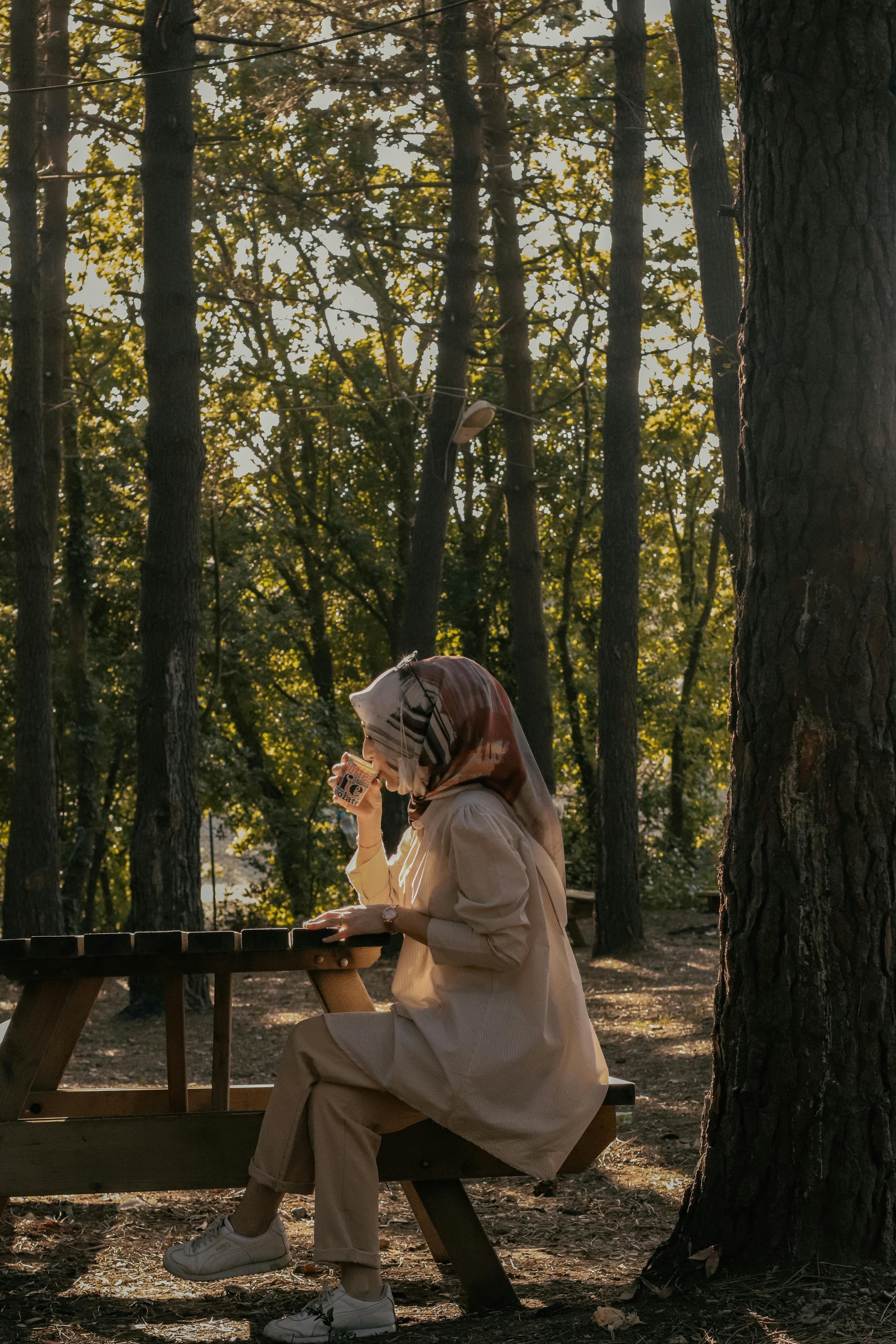 A woman sitting alone at a picnic table in a forest park, sipping coffee.