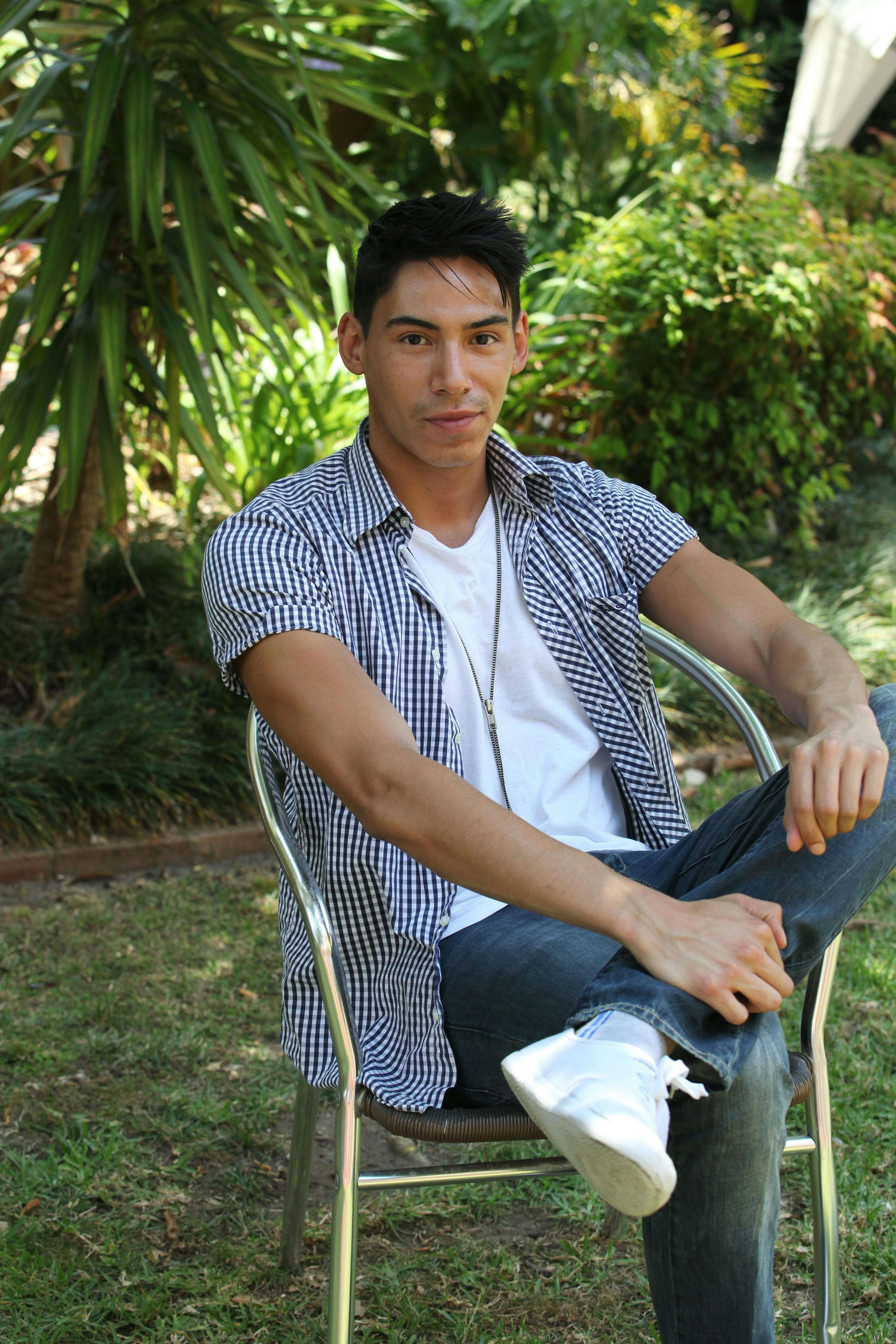 A young man in a checkered shirt sits outdoors, showcasing a relaxed and confident pose.