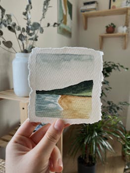 A hand holding a watercolor painting of a peaceful landscape in a cozy interior.