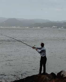 Side view of unrecognizable male in casual clothes with fishing rod standing on rocky coast and catching fish in rippling water in gloomy day