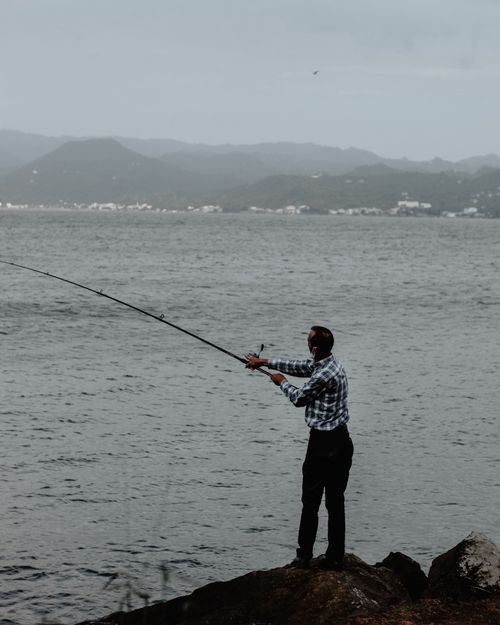 Free Side view of unrecognizable male in casual clothes with fishing rod standing on rocky coast and catching fish in rippling water in gloomy day Stock Photo