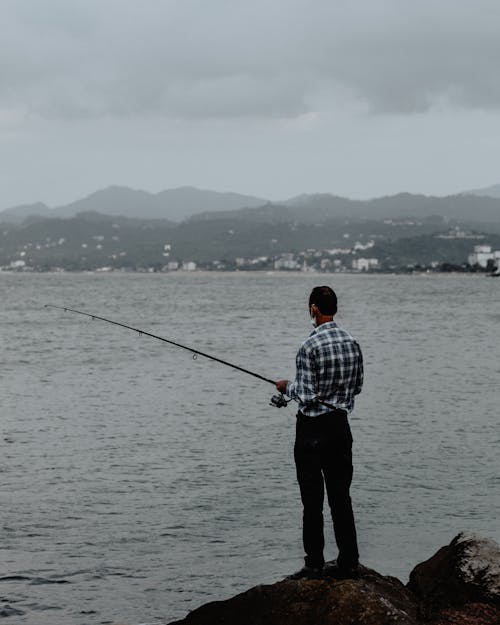 Back view of unrecognizable fisherman standing on rocky coast near rippling water and catching fish with fishing rod against view of mountains in cloudy gloomy day