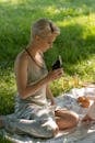 Woman in White Tank Top and Gray Pants Sitting on Green Grass Field Holding Black Smartphone