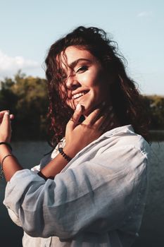 A woman in white long sleeves poses confidently by a serene lake, smiling warmly on a sunny day.