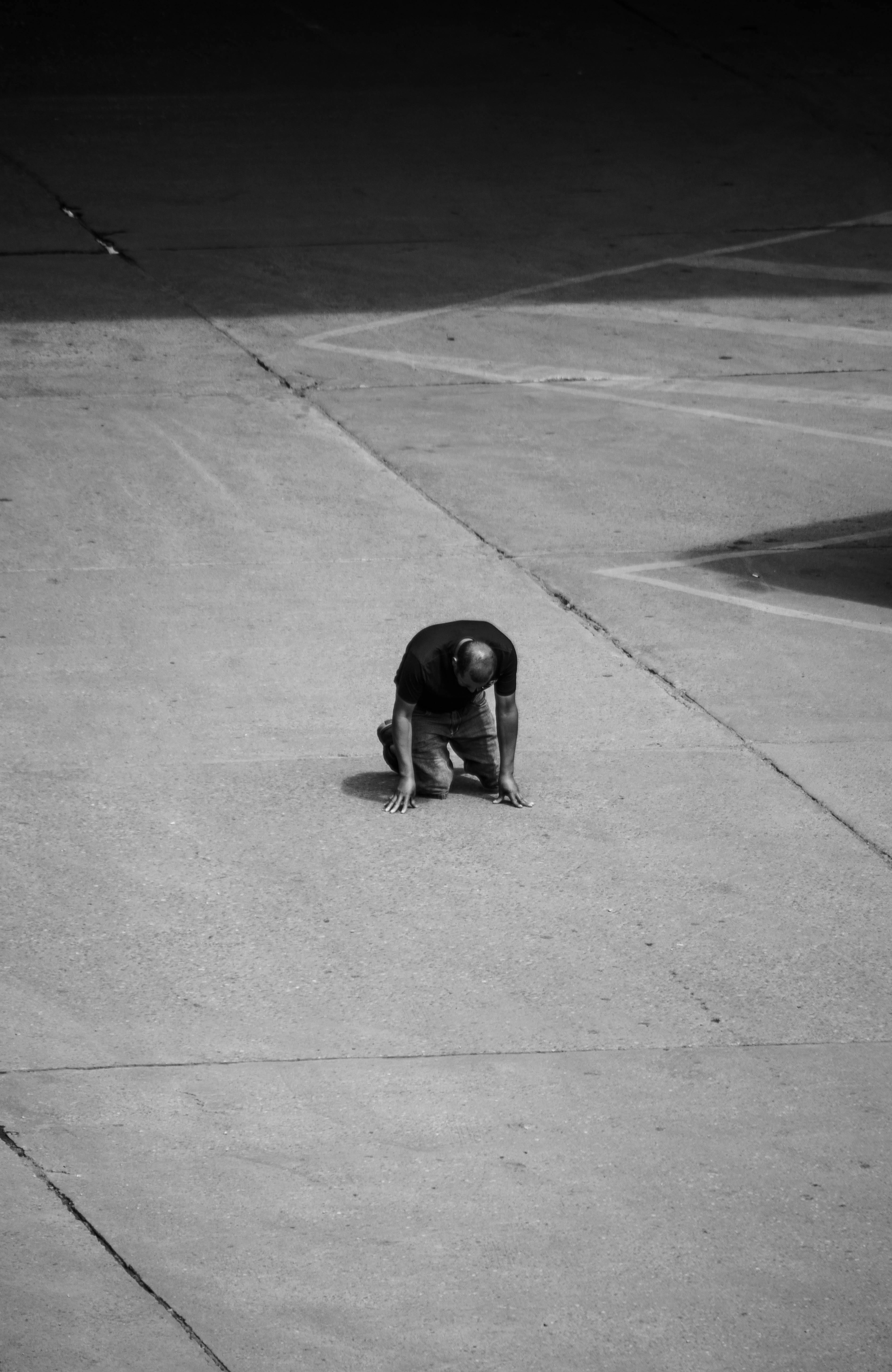 Monochrome image of a man kneeling on empty concrete pavement, emphasizing solitude.