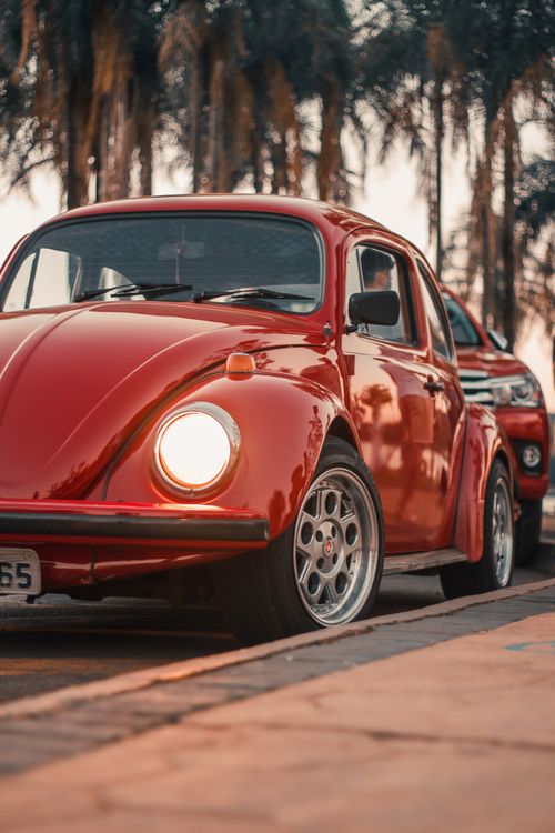Free Close-up of a classic red Volkswagen Beetle parked outdoors against a palm tree backdrop at sunset. Stock Photo