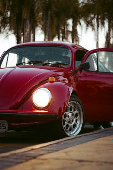 A vintage red Volkswagen Beetle with headlights on, parked alongside a road.