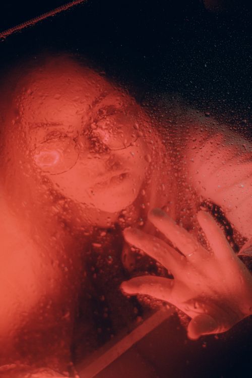 Free Artistic portrait of a woman with droplets on glass, illuminated by dramatic red lighting. Stock Photo