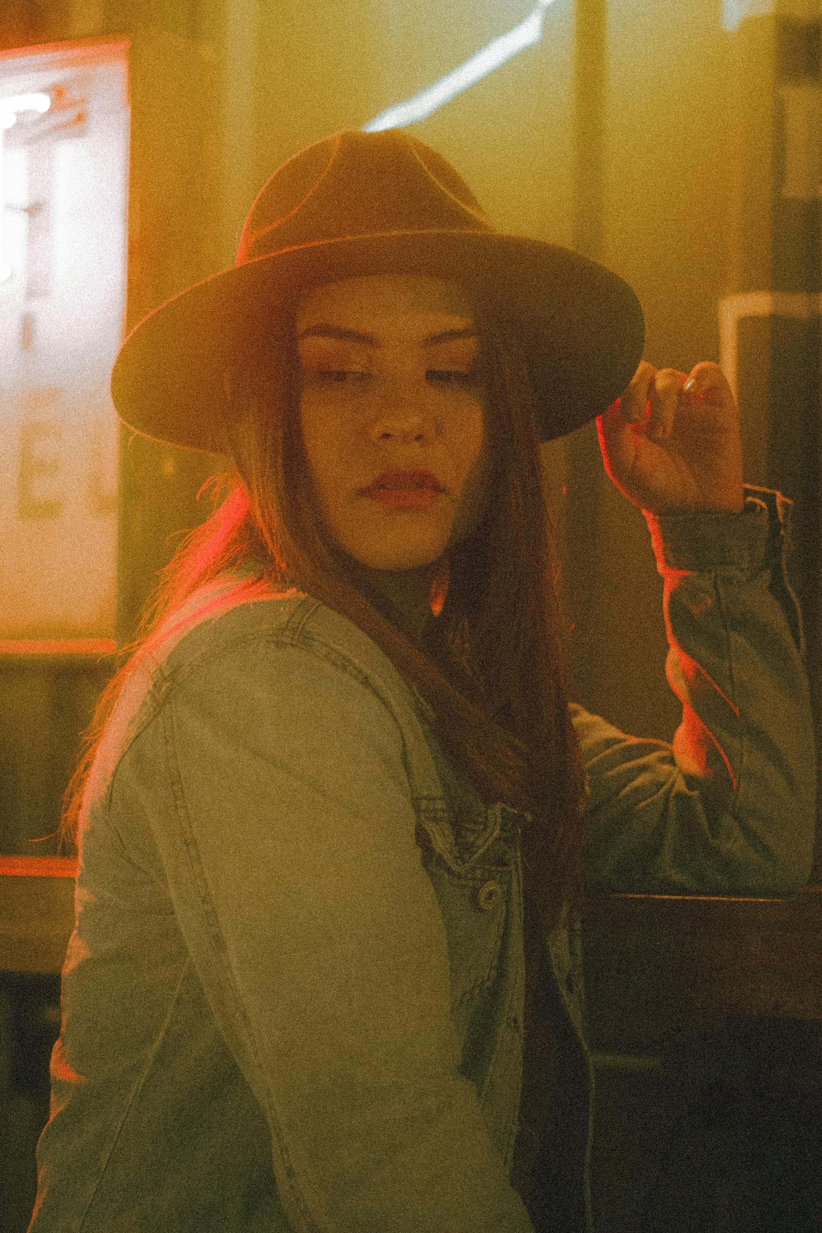 A contemplative woman in a denim jacket and hat under warm neon lighting, exuding a moody and nostalgic vibe.