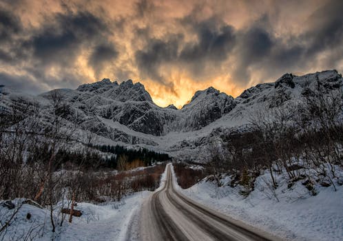 A scenic road leads through snowy peaks at sunrise in Flakstad, Norway, capturing the tranquil beauty of a winter landscape.