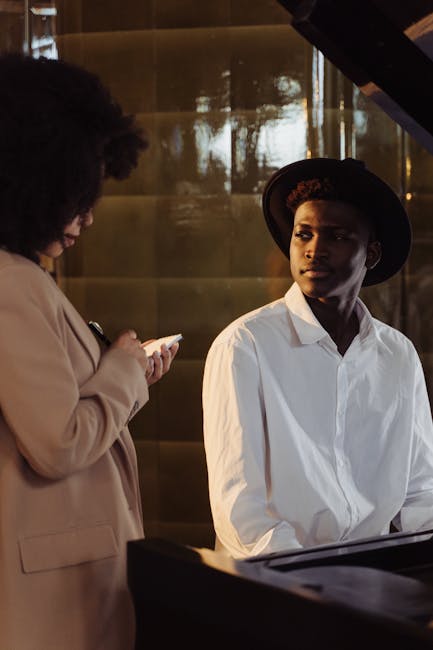 A musician in a hat sits at a piano while a woman takes notes during an interview indoors.
