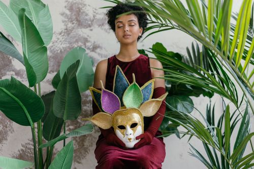 Free A woman dressed elegantly in a deep maroon dress with a colorful carnival mask amidst lush greenery. Stock Photo