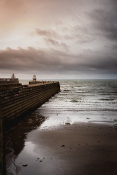 Moody seascape featuring a pier and seawall with a dramatic sunset over the ocean.