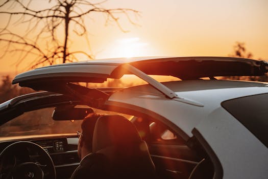 Person in convertible car driving during sunset with sun glare creating a warm atmosphere.