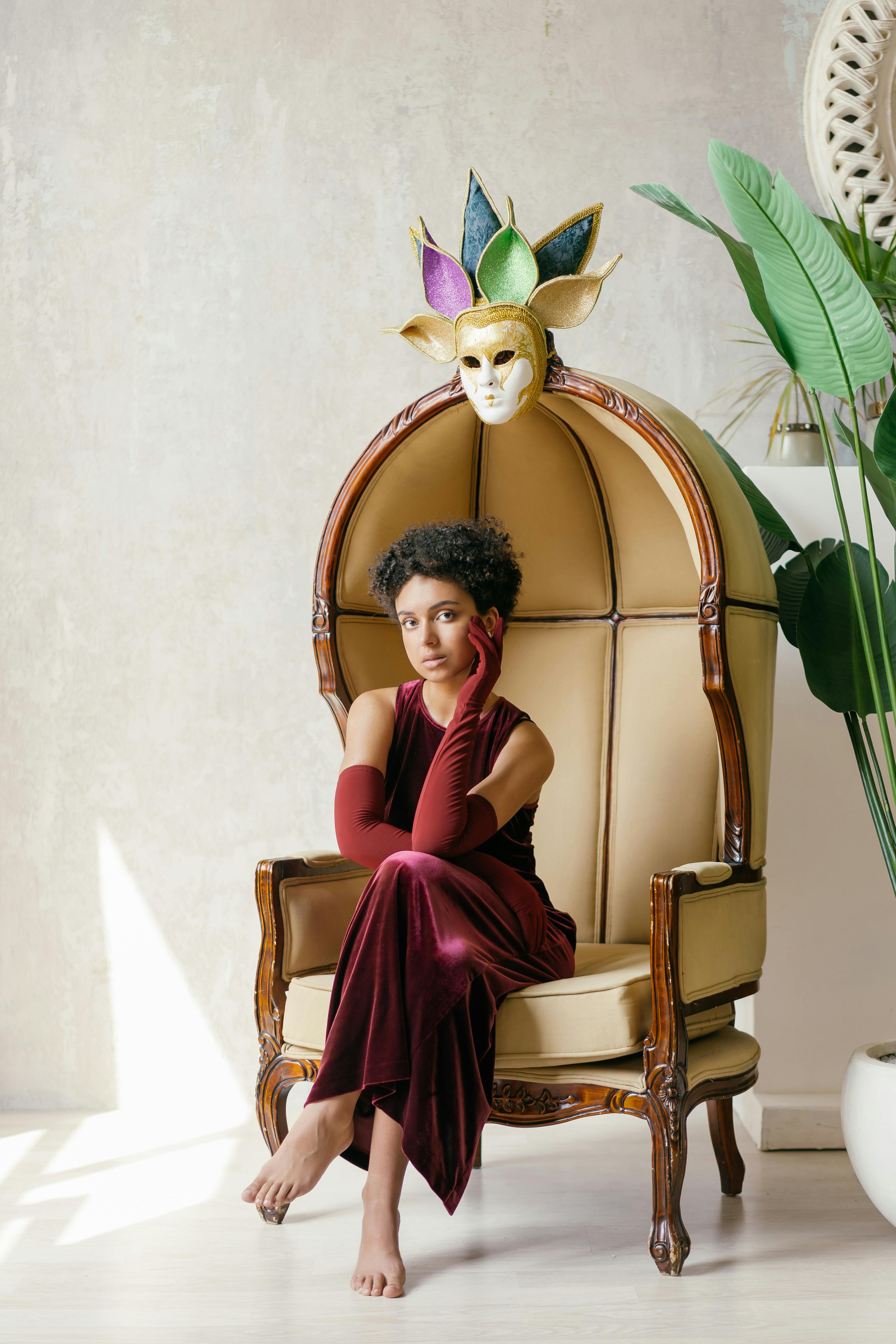 Free A woman in a maroon dress sits elegantly with a masquerade mask overhead, surrounded by plants. Stock Photo
