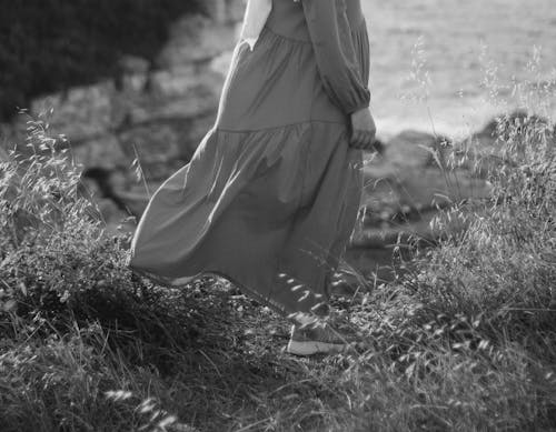 Grayscale Photo of a Woman in a Dress Standing on Grass Covered Ground