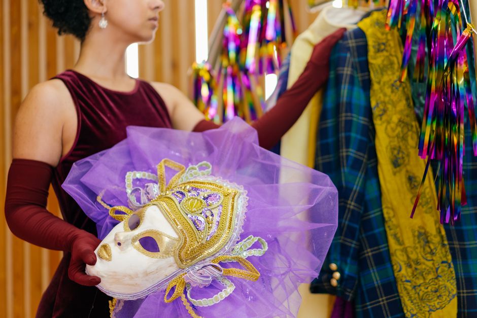 The Masquerade Ball 2 A woman elegantly holds a decorative masquerade mask beside vibrant, colorful costumes. | BocaVibe