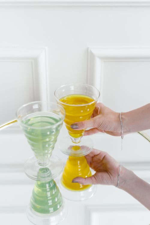 Free Vibrant green and yellow cocktails reflecting on a glass table with elegant hands holding them. Stock Photo