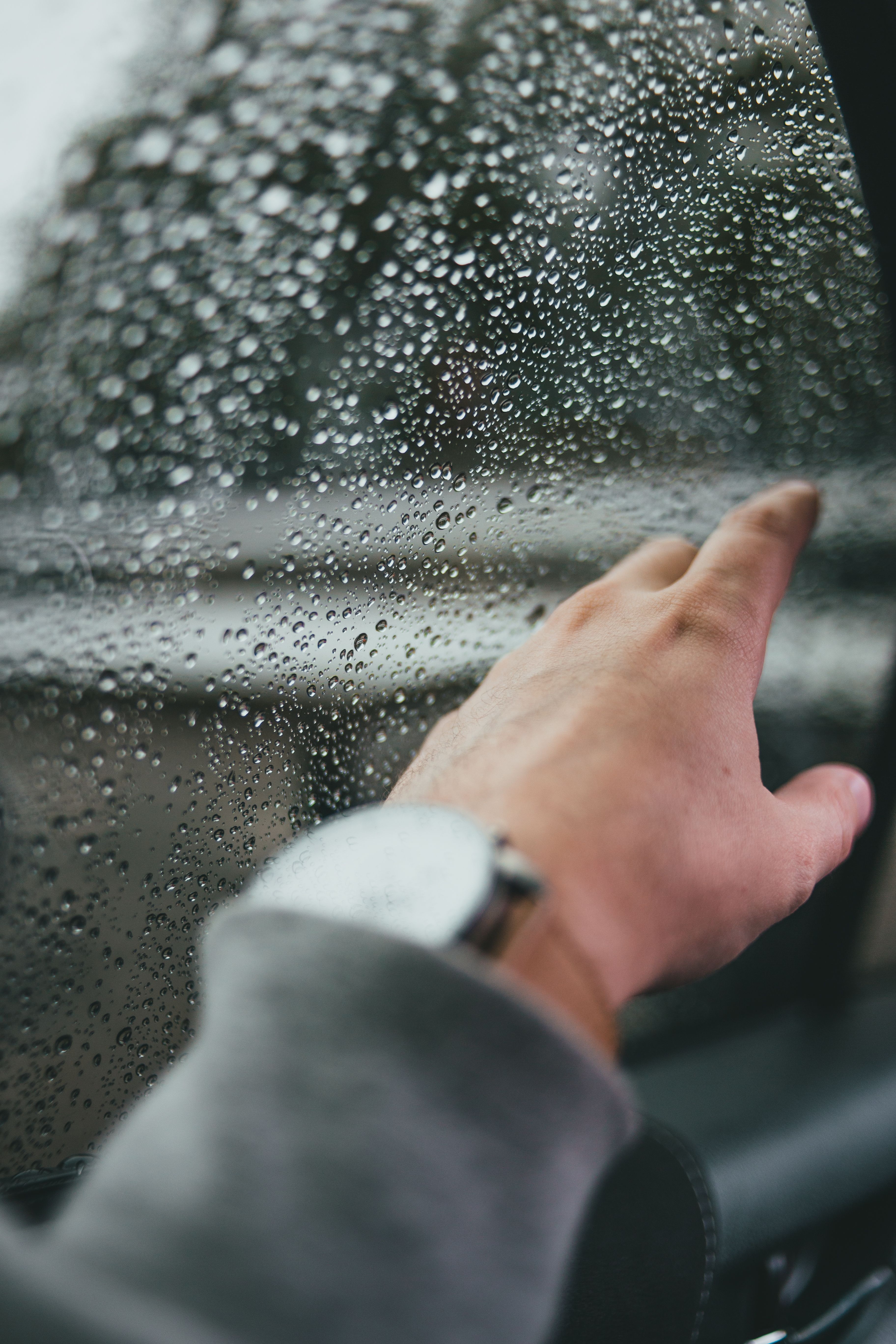 Close-up of a hand with a watch, touching raindrop-covered car window.