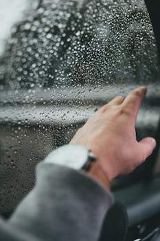 Close-up of a hand with a watch, touching raindrop-covered car window.